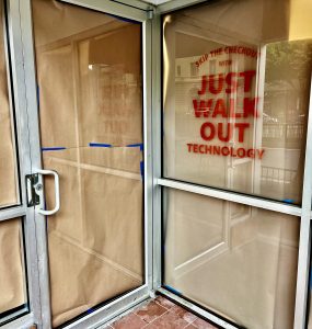 An image of two glass doors which intersect at a corner; the door on the right displays large orange text, "skip the checkout with Just Walk Out technology"