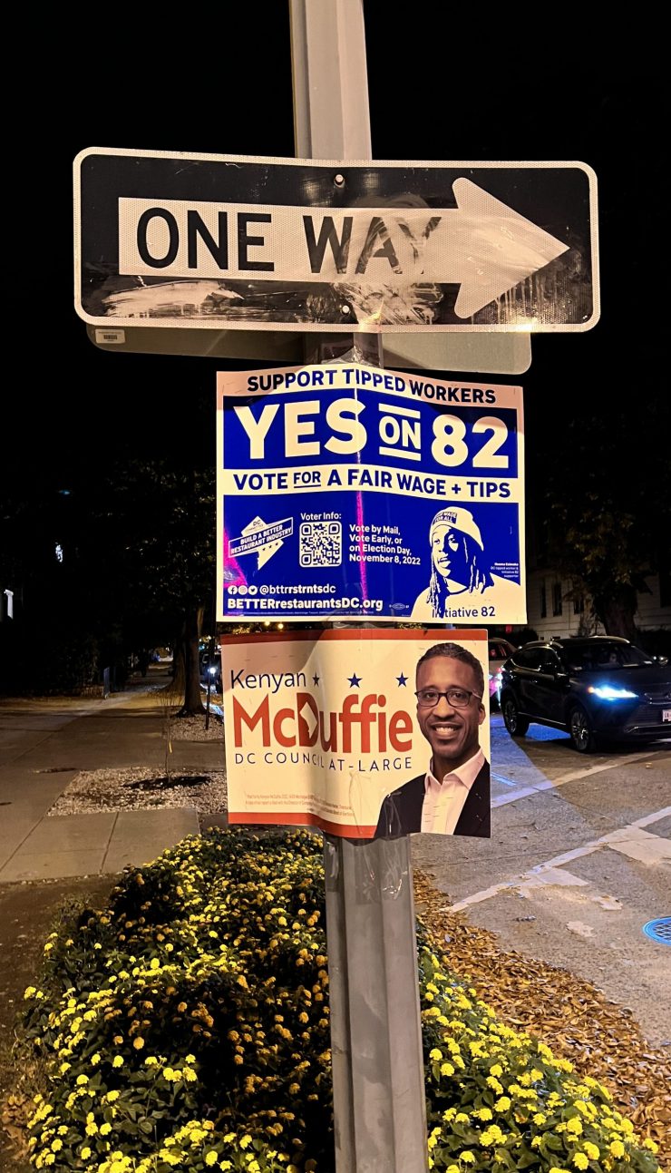 a pole on a street corner at night with three signs. a scuffed One Way sign points to the right; below it are two campaign signs; the top campaign sign is blue and white and says "Yes on 82" and voting information; the second campaign sign is for Kenyan McDuffie, D.C. Council at Large