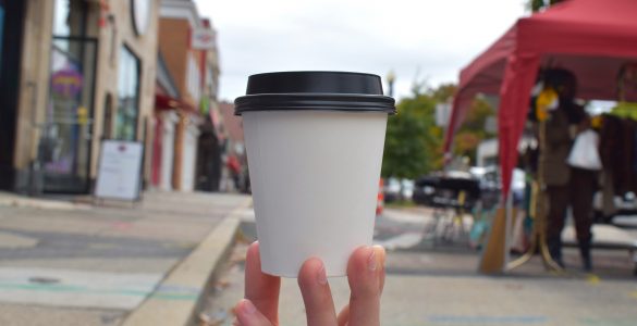 An unmarked to-go coffee cup is held up with a blurred background of the Cleveland Park main street