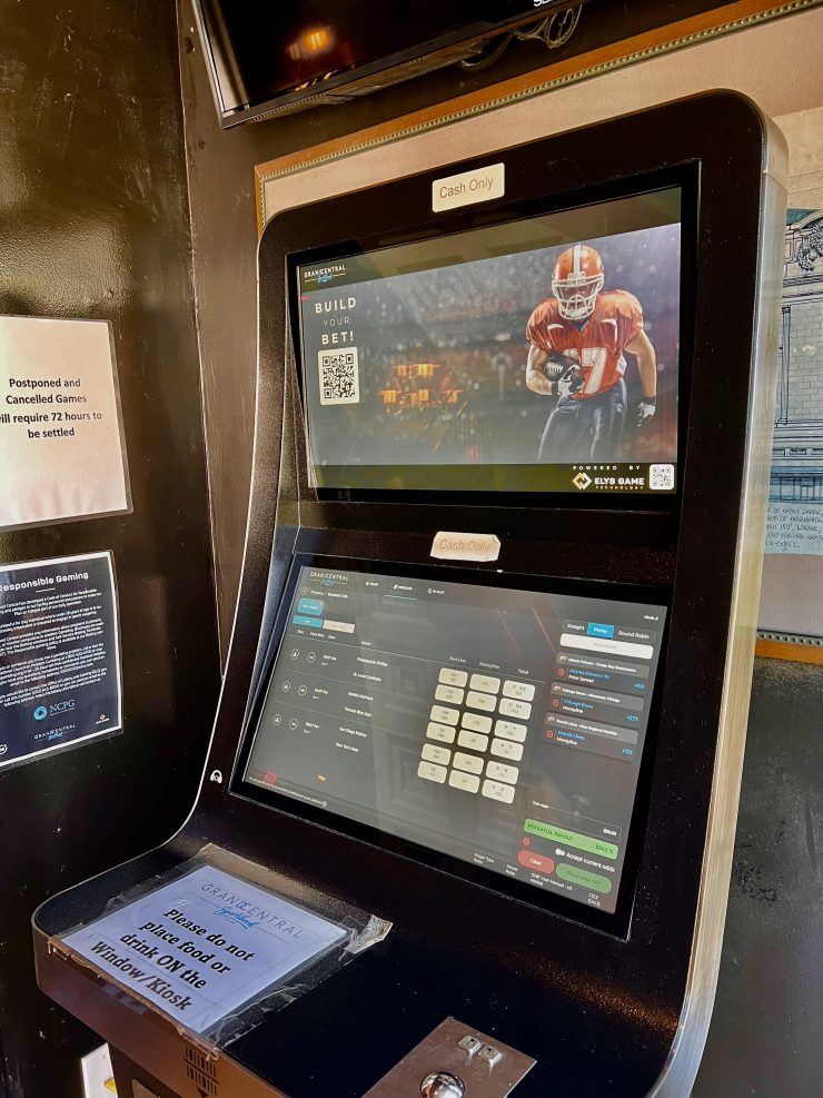 the top half of a sports betting kiosk at 3/4 view with two horizontal screens. the top screen shows a football player and a QR code to scan to "build your bet." the bottom touch screen shows betting details