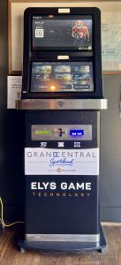 front view of a black and silver sports betting kiosk. the top half has two horizontal screens. the bottom half has a barcode scanner, credit card and cash readers and a ticket slot. below are logos for Grand Central and Elys Game Technology