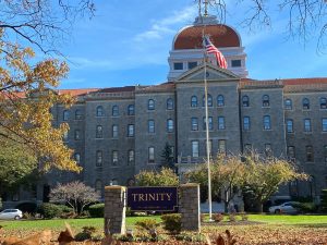A sign for Trinity Washington University sits in front of a large building, the campus Main Hall.