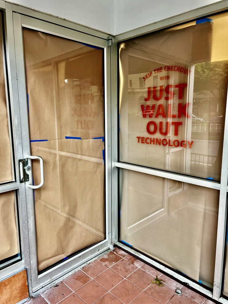 An image of two glass doors which intersect at a corner; the door on the right displays large orange text, "skip the checkout with Just Walk Out technology"
