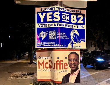 a pole on a street corner at night with three signs. a scuffed One Way sign points to the right; below it are two campaign signs; the top campaign sign is blue and white and says "Yes on 82" and voting information; the second campaign sign is for Kenyan McDuffie, D.C. Council at Large