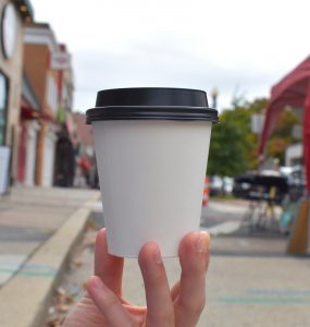 An unmarked to-go coffee cup is held up with a blurred background of the Cleveland Park main street