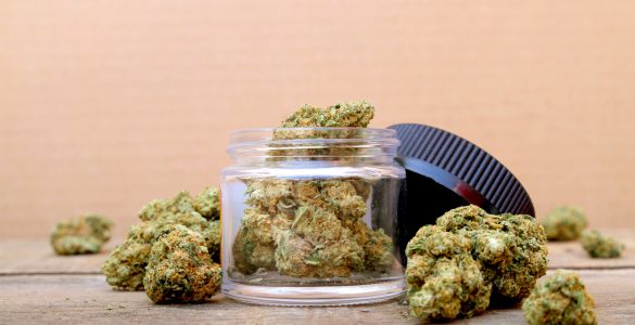 An open clear jar with marijuana buds inside, surrounded by more buds outside.