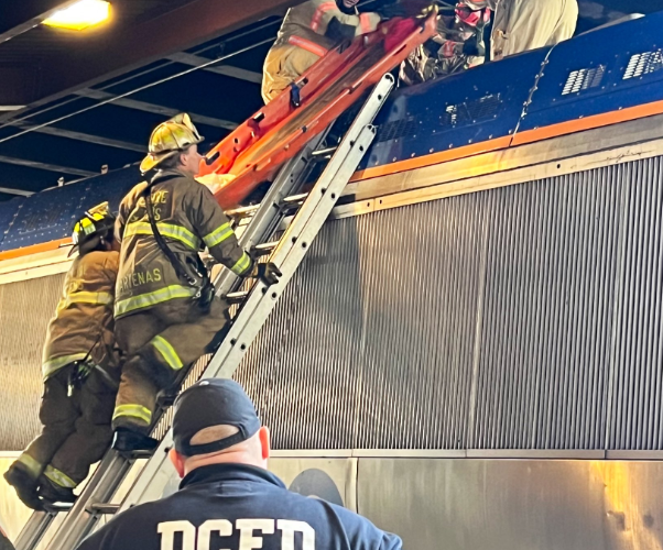 Firefighters climb a ladder on top of a rail train to assist a man offscreen.