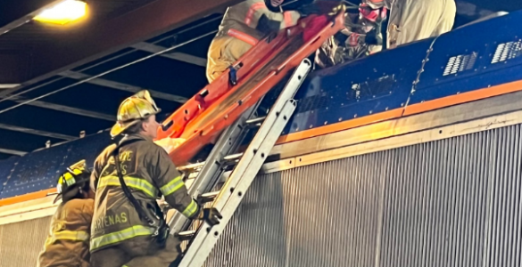 Firefighters climb a ladder on top of a rail train to assist a man offscreen.