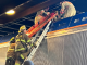 Firefighters climb a ladder on top of a rail train to assist a man offscreen.