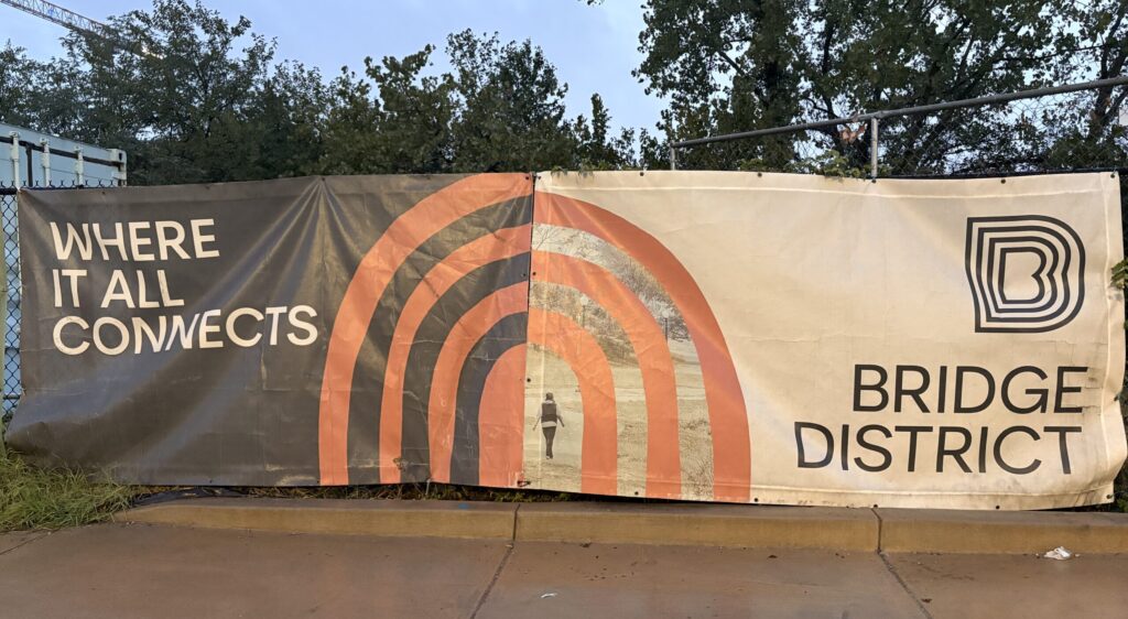 A perforated vinyl mesh banner reads "WHERE IT ALL CONNECTS" on its left side in white lettering and a graphite background. "BRIDGE DISTRICT" in graphite lettering and a white background is displayed on the right of the banner. An interlocking, seven-tiered orange arch sits at the center of the banner. The arches are interwoven with a photo of an individual walking through a grassy field. 