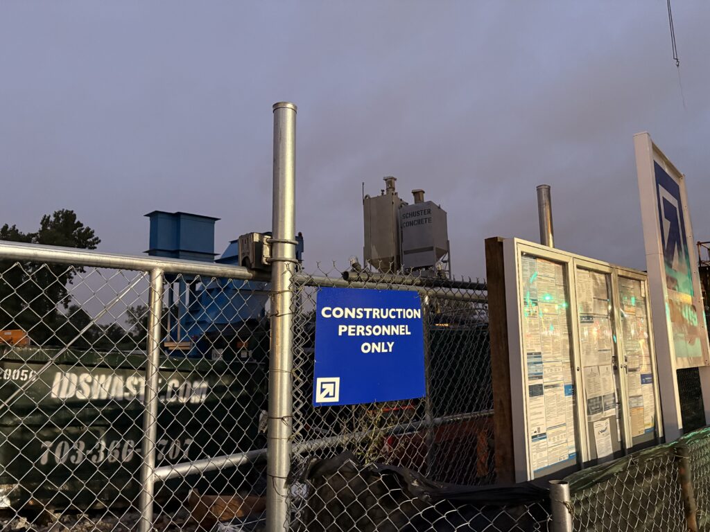 Under a darkening evening sky, a royal blue sign reads "Construction Personnel Only" in white lettering on a steel fence. Beside it, there are three permit posting boxes and a large sign directing visitors to the right towards the Anacostia metro. A dumpster and construction materials lie behind the fence. 