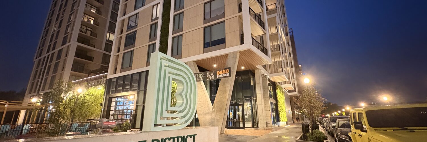 A tan, concrete monument sign reads "BRIDGE DISTRICT LIVING" in forest green lettering. The Bridge District logo, a series of green, concentric letter Ds, sits atop it. A high rise apartment complex towers behind the sign.