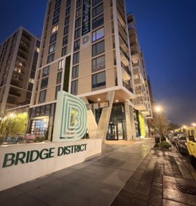 A tan, concrete monument sign reads "BRIDGE DISTRICT LIVING" in forest green lettering. The Bridge District logo, a series of green, concentric letter Ds, sits atop it. A high rise apartment complex towers behind the sign.