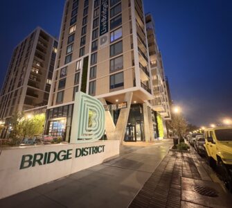 A tan, concrete monument sign reads "BRIDGE DISTRICT LIVING" in forest green lettering. The Bridge District logo, a series of green, concentric letter Ds, sits atop it. A high rise apartment complex towers behind the sign.