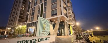 A tan, concrete monument sign reads "BRIDGE DISTRICT LIVING" in forest green lettering. The Bridge District logo, a series of green, concentric letter Ds, sits atop it. A high rise apartment complex towers behind the sign.