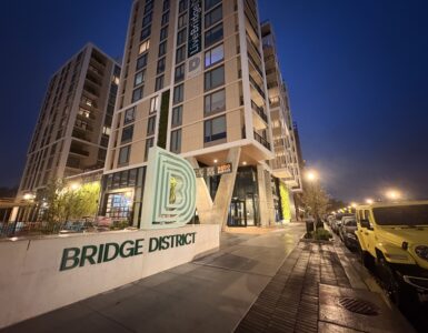 A tan, concrete monument sign reads "BRIDGE DISTRICT LIVING" in forest green lettering. The Bridge District logo, a series of green, concentric letter Ds, sits atop it. A high rise apartment complex towers behind the sign.