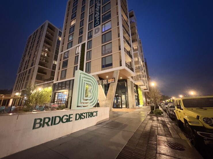 A tan, concrete monument sign reads "BRIDGE DISTRICT LIVING" in forest green lettering. The Bridge District logo, a series of green, concentric letter Ds, sits atop it. A high rise apartment complex towers behind the sign.
