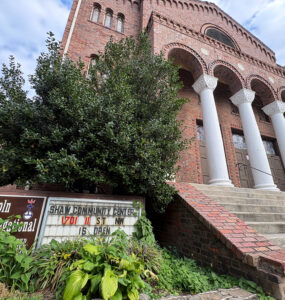Looking up at the historic Lincoln Temple, an overgrown sign reads "Lincoln Congregational Temple United Church of Christ" and "Shaw Community Center 1701 11 St NW is open."
