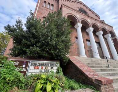 Looking up at the historic Lincoln Temple, an overgrown sign reads "Lincoln Congregational Temple United Church of Christ" and "Shaw Community Center 1701 11 St NW is open."