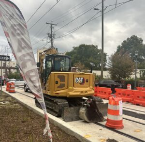 A yellow construction vehicle sits in the middle of laid light rail tracks between orange traffic cones and a flag on the sidewalk that says "Barber."