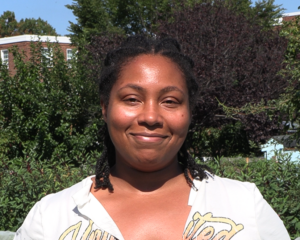 Joya Wade smiles at the camera in front of bushes and trees in this headshot.