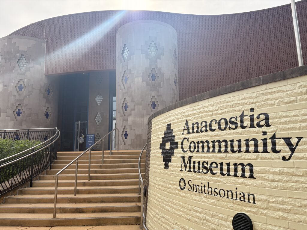 A tan, brick monument sign reads "Anacostia Community Museum" with "Smithsonian" below it. A black, diamond-shaped emblem is painted to the left of the words. A stairway leads upward to a darker-bricked building with tribal-patterned columns flanking its entryway. 
