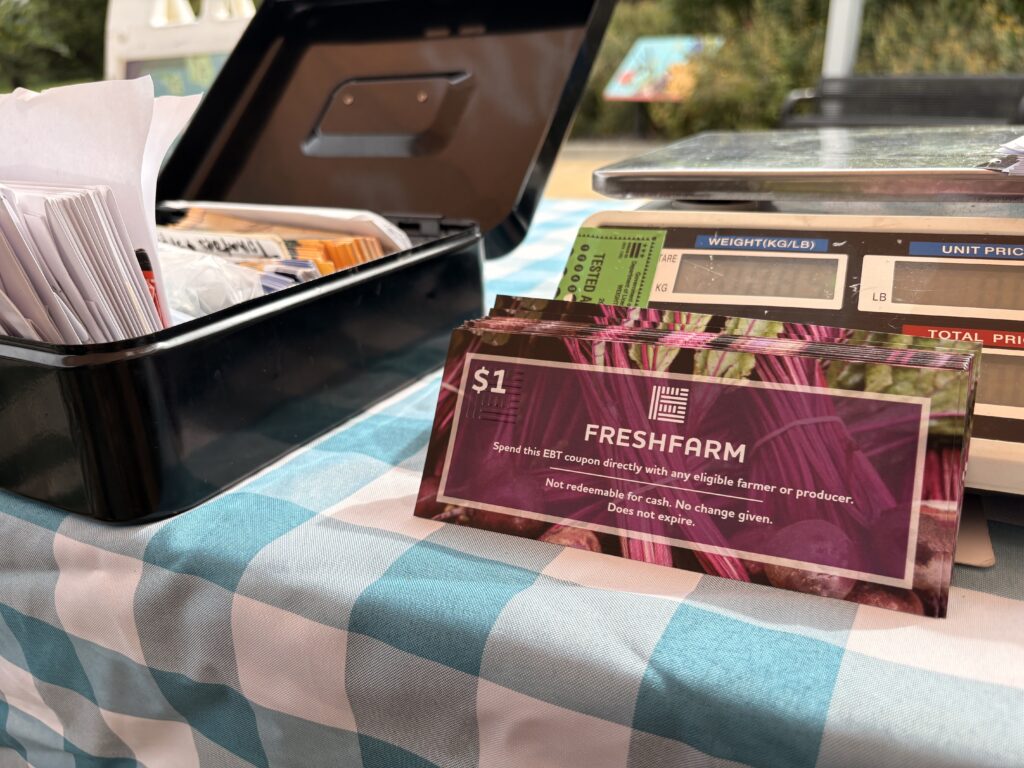 A stack of plum-colored FRESHFARM coupons rest against a food scale. The scale sits next to a lockbox filled with with envelopes and other items. The coupon, scale, and lockbox all sit atop a teal and white gingham table cloth. 