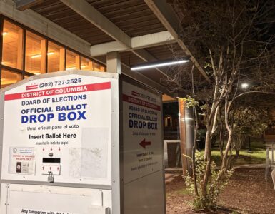 A white, steel box with blue and red lettering has a sign that reads "District of Columbia," "Board of Elections," and "Official Ballot Drop Box," each on individual lines. Arrows point to where ballots can be dropped into the box.